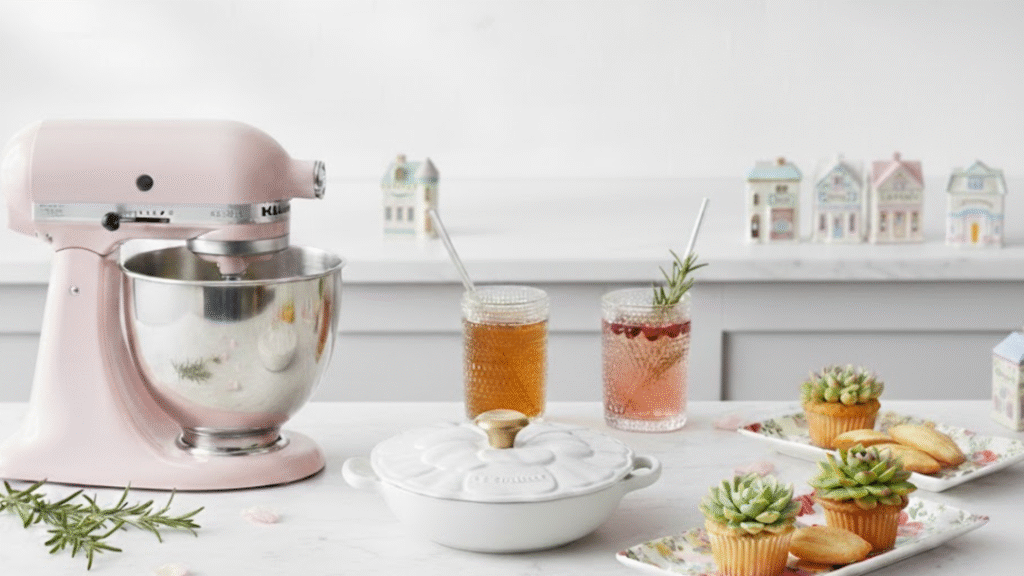 A stylized kitchen scene inspired by the products in Chapter 1 of the holiday wishbook. A soft pink mixer, vintage glasses containing festive refreshments, ceramic spice houses dispered throughout the countertop, a white floral braiser, and sweet cupcake treats arranged on floral serving trays.