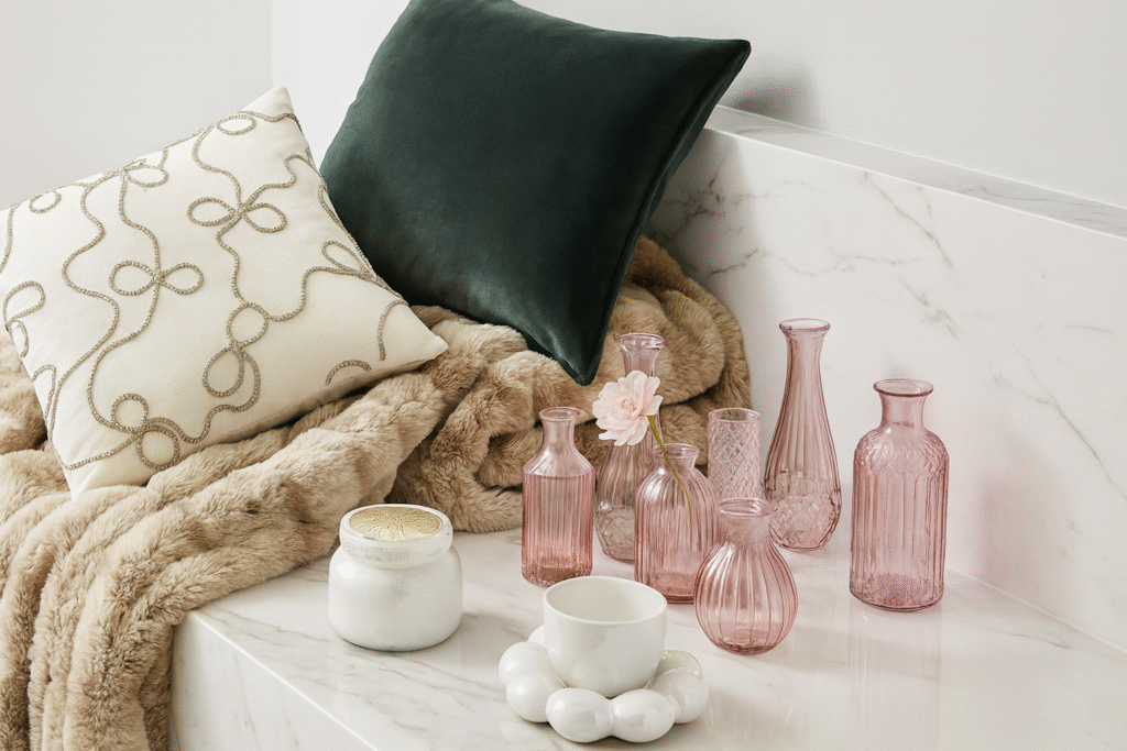 A stylized scene featuring items inspired by chapter 2 of the holiday wishbook. A tan faux fur blanket draped on a marble counter with antique rose bud vases, a velvet spruce green pillow, a cream accent pillow with sparkling bow details, a pearlescent cup on a cloud saucer, and a candle.